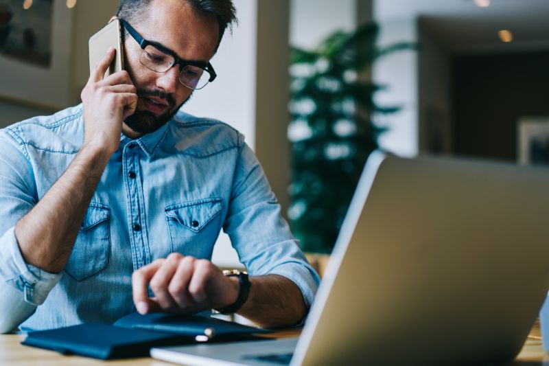 Man checking watch while on phone with laptop open, waiting for IRS tax refund approval status