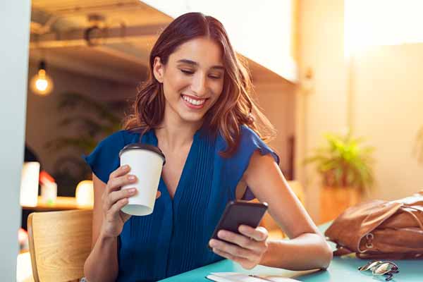 Woman smiling at her phone while enjoying coffee, reading about smart ways to reduce capital gains taxes and build wealth.