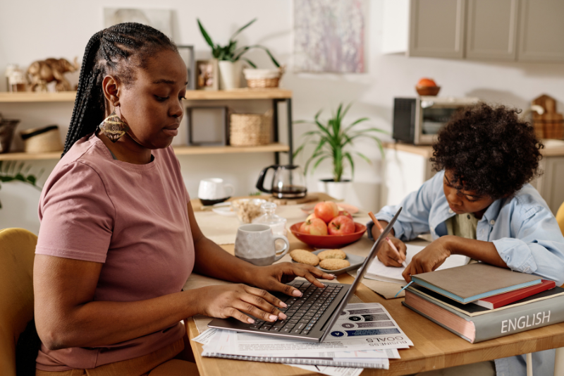 A mom working on her laptop while her son does his homework at the kitchen table, symbolizing the balance of work and family life for low-income families seeking government assistance programs.