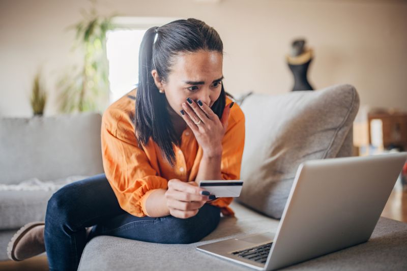 Young woman sitting on couch, distressed while looking at her laptop, holding a credit card, symbolizing the stress of high-interest loans and debt traps.