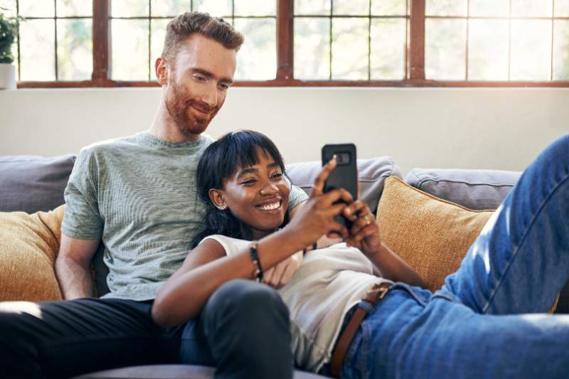 Husband and wife relaxing on couch looking at phone, researching if a spouse can be claimed as a dependent on taxes.