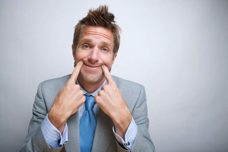 Man forcing a smile with his fingers, illustrating toxic positivity in the workplace and the pressure to appear happy at work.